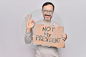 Middle age handsome man on disagreement holding banner with not my president message doing ok sign with fingers, smiling friendly