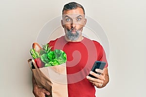 Middle age handsome man holding bag of groceries using smartphone making fish face with mouth and squinting eyes, crazy and
