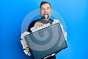 Middle age handsome man holding briefcase full of dollars sticking tongue out happy with funny expression