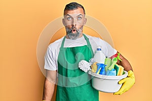 Middle age handsome man holding cleaning products scared and amazed with open mouth for surprise, disbelief face