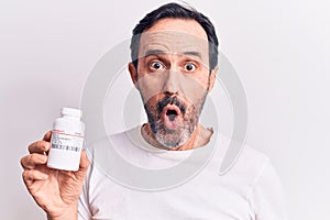 Middle age handsome man holding jar of pills standing over isolated white background scared and amazed with open mouth for