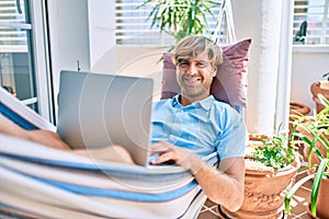 Middle age handsome man at the terrace of his house relaxing lying on a hammock working with laptop