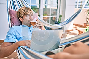 Middle age handsome man at the terrace of his house relaxing lying on a hammock working with laptop and drinking a fresh beer