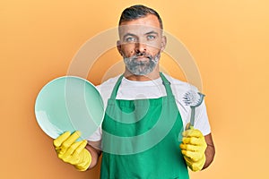 Middle age handsome man wearing apron holding scourer washing dishes relaxed with serious expression on face