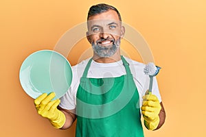 Middle age handsome man wearing apron holding scourer washing dishes smiling with a happy and cool smile on face