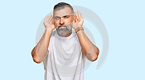 Middle age handsome man wearing casual white tshirt trying to hear both hands on ear gesture, curious for gossip