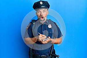 Middle age handsome man wearing police uniform writing traffic fine afraid and shocked with surprise and amazed expression, fear