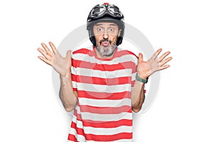 Middle age handsome motorcyclist man wearing moto helmet over isolated white background clueless and confused with open arms, no