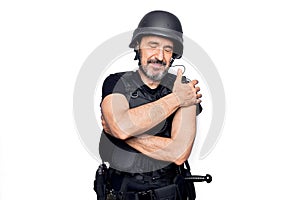 Middle age handsome policeman wearing police bulletproof vest and security helmet hugging oneself happy and positive, smiling