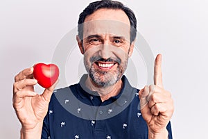 Middle age handsome romantic man holding plastic heart over isolated white background smiling with an idea or question pointing