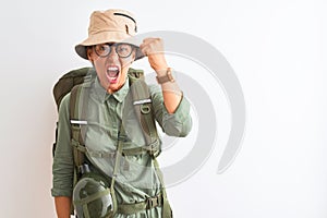 Middle age hiker woman wearing backpack canteen hat glasses over isolated white background angry and mad raising fist frustrated