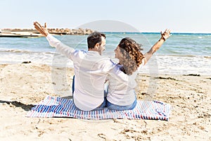 Middle age hispanic couple on back view sitting on the towel hugging at the beach