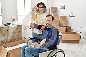 Middle age hispanic couple and dog sitting on wheelchair at new home with angry face, negative sign showing dislike with thumbs