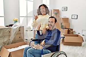 Middle age hispanic couple and dog sitting on wheelchair at new home pointing thumb up to the side smiling happy with open mouth