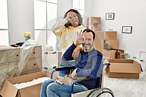 Middle age hispanic couple and dog sitting on wheelchair at new home smiling happy doing ok sign with hand on eye looking through