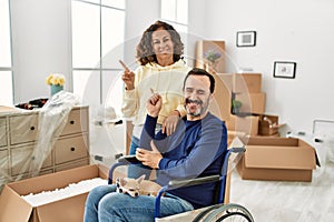 Middle age hispanic couple and dog sitting on wheelchair at new home smiling happy pointing with hand and finger to the side