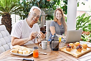 Middle age hispanic couple having breakfast using smartphone and laptop at the terrace