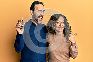 Middle age hispanic couple holding keys of new home pointing thumb up to the side smiling happy with open mouth