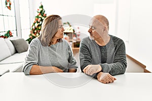 Middle age hispanic couple sitting on the table by christmas tree looking to side, relax profile pose with natural face with