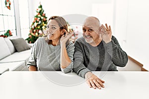 Middle age hispanic couple sitting on the table by christmas tree smiling with hand over ear listening an hearing to rumor or