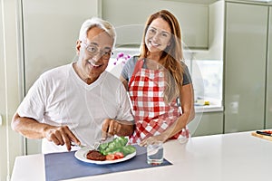 Middle age hispanic couple smiling happy eating beef with salad at the kitchen