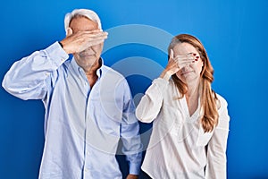 Middle age hispanic couple standing over blue background covering eyes with hand, looking serious and sad