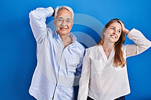 Middle age hispanic couple standing over blue background smiling confident touching hair with hand up gesture, posing attractive