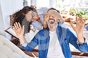 Middle age hispanic couple surprise with hands on eyes sitting on hammock at terrace