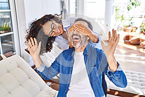 Middle age hispanic couple surprise with hands on eyes sitting on hammock at terrace