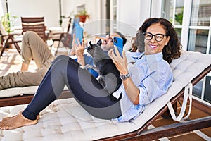 Middle age hispanic couple using smartphone and touchpad lying on hammock with dog at terrace