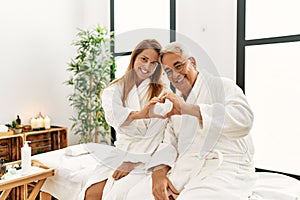 Middle age hispanic couple wearing bathrobe at wellness spa smiling in love showing heart symbol and shape with hands