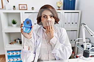 Middle age hispanic dentist woman working at dentist clinic holding invisible aligner covering mouth with hand, shocked and afraid