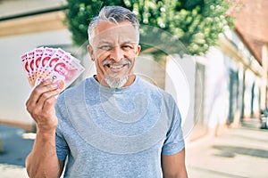 Middle age hispanic grey-haired man smiling happy holding chinese yuan banknotes at the city
