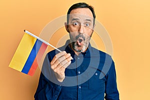 Middle age hispanic man holding colombia flag scared and amazed with open mouth for surprise, disbelief face