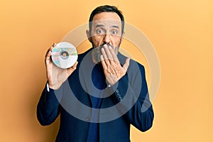 Middle age hispanic man holding compact disc covering mouth with hand, shocked and afraid for mistake