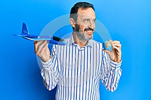 Middle age hispanic man holding plane toy and world ball smiling looking to the side and staring away thinking