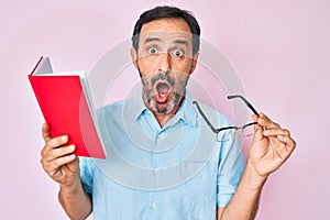 Middle age hispanic man reading book holding glasses afraid and shocked with surprise and amazed expression, fear and excited face