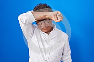 Middle age hispanic man standing over blue background covering eyes with arm, looking serious and sad