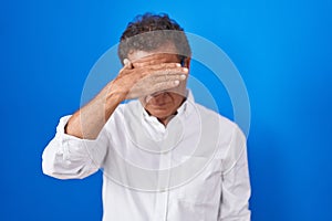 Middle age hispanic man standing over blue background covering eyes with hand, looking serious and sad