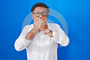 Middle age hispanic man standing over blue background shocked covering mouth with hands for mistake