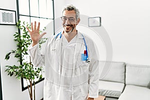 Middle age hispanic man wearing doctor uniform and stethoscope at waiting room showing and pointing up with fingers number five