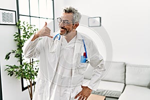 Middle age hispanic man wearing doctor uniform and stethoscope at waiting room smiling doing phone gesture with hand and fingers
