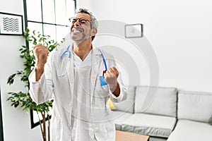 Middle age hispanic man wearing doctor uniform and stethoscope at waiting room very happy and excited doing winner gesture with