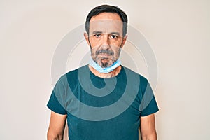 Middle age hispanic man wearing medical mask on neck thinking attitude and sober expression looking self confident