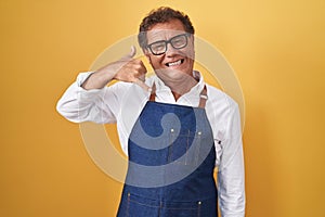 Middle age hispanic man wearing professional cook apron smiling doing phone gesture with hand and fingers like talking on the