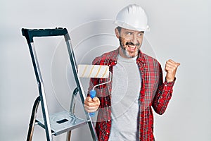 Middle age hispanic painter man holding roller using ladder pointing thumb up to the side smiling happy with open mouth