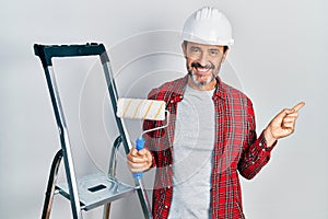 Middle age hispanic painter man holding roller using ladder smiling happy pointing with hand and finger to the side
