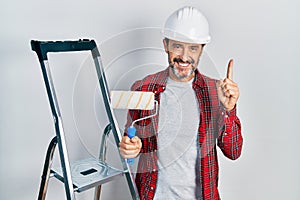 Middle age hispanic painter man holding roller using ladder surprised with an idea or question pointing finger with happy face,