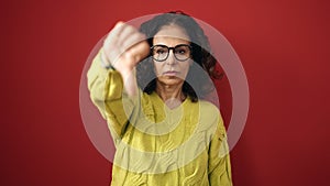 Middle age hispanic woman doing negative gesture with thumb down over isolated red background