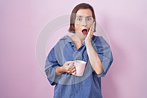 Middle age hispanic woman drinking a cup coffee afraid and shocked, surprise and amazed expression with hands on face